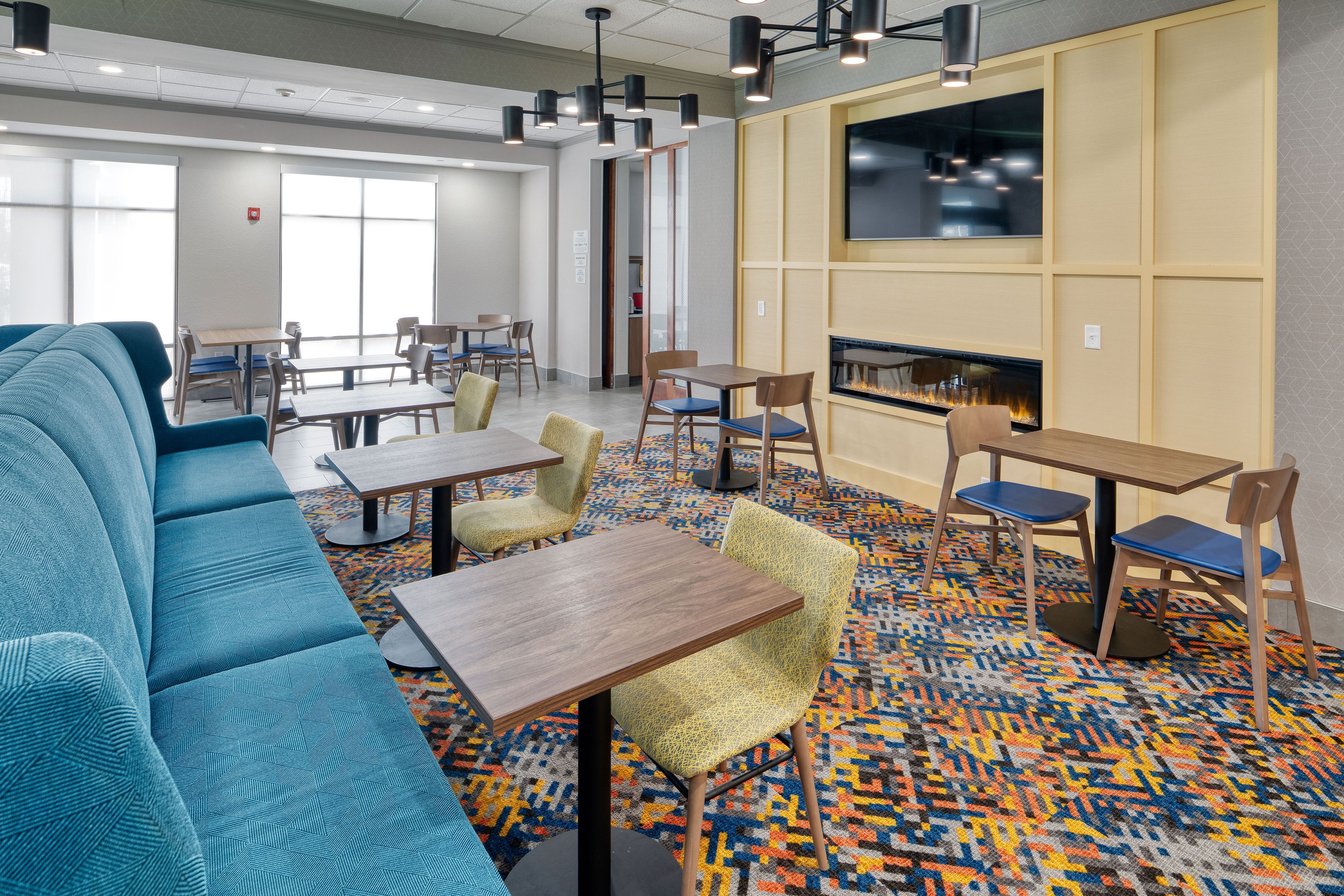 A hotel lobby with a blue sofa, wooden tables, chairs, a patterned carpet, a fireplace, and a television on the wall.