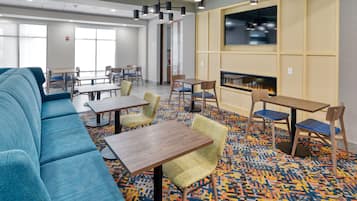 A hotel lobby with a blue sofa, wooden tables, chairs, a patterned carpet, a fireplace, and a television on the wall.