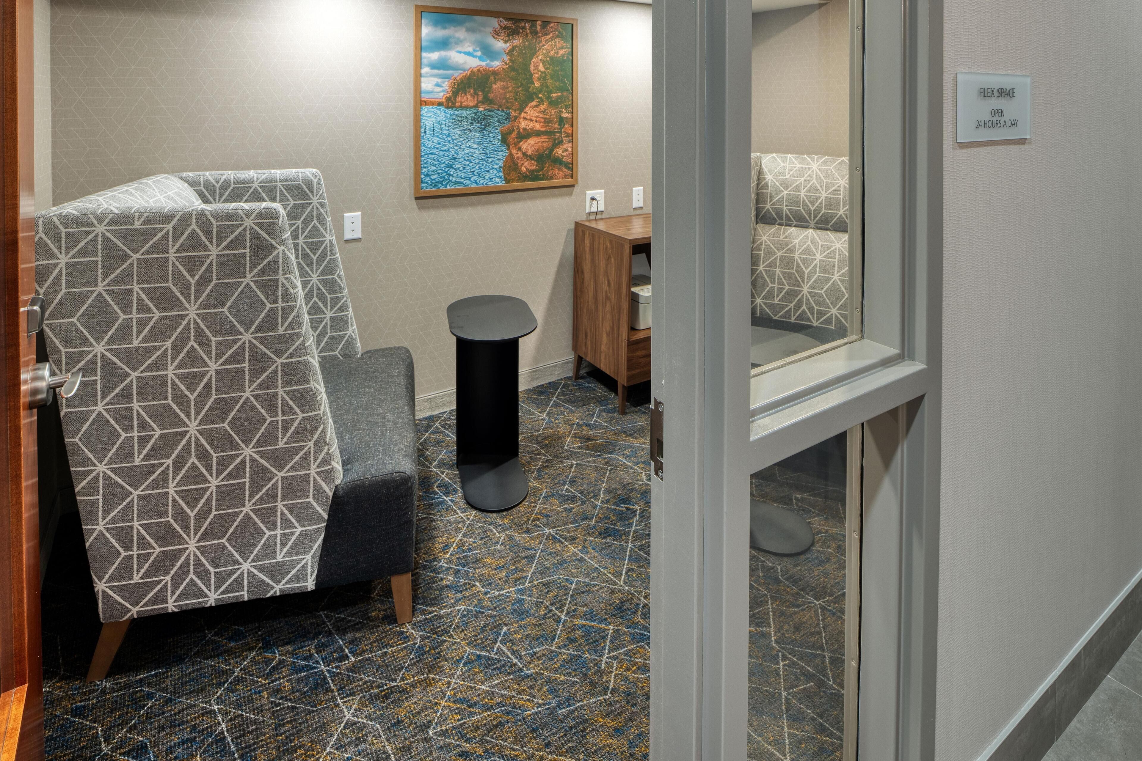 A hotel room with a patterned chair, a small round table, a painting on the wall, and a sign indicating the room is a Flex Space open 24 hours.