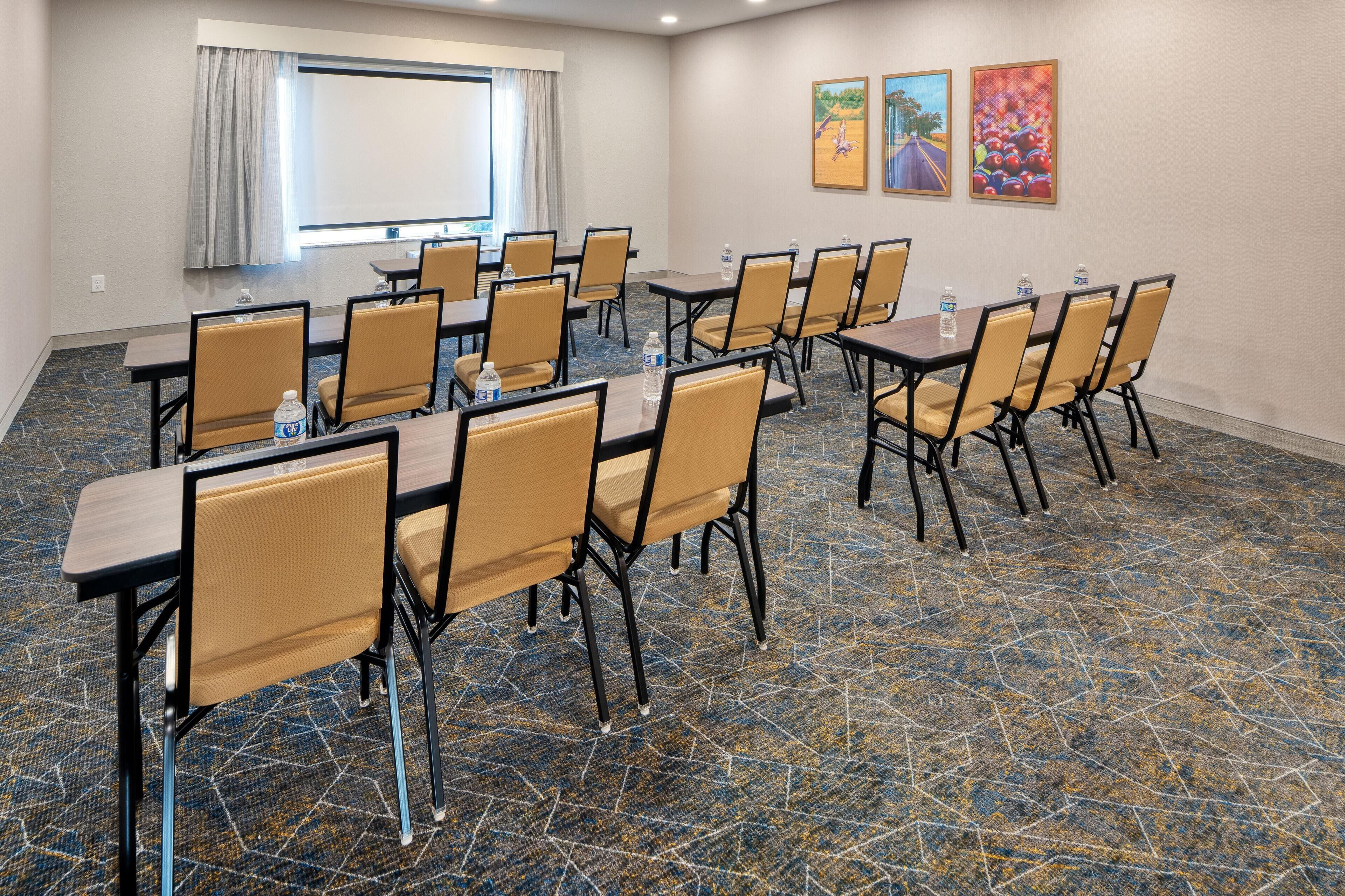 Une salle de conférence équipée de tables et de chaises, d’un écran de projection et d’une fenêtre avec des rideaux.
