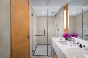 A modern bathroom with a glass shower enclosure, a wooden door, a white sink with a mirror, and a vase with purple flowers.