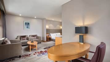 Habitación de hotel con sofá, una mesa redonda de madera, una silla, cama con mesita de noche y lámpara.