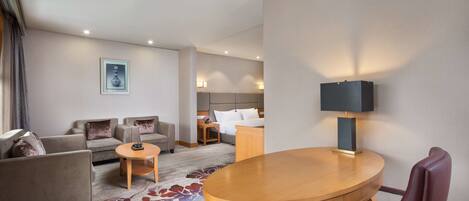 A hotel room with a sofa set, a round wooden table, a single chair, a bed with a nightstand, and a lamp.