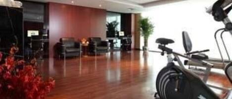 A modern hotel lobby with a wooden floor, a reception desk, seating area, and exercise equipment.