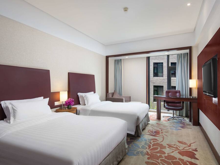 A hotel room with two beds, a desk, a chair, a TV, and a view of the outside.