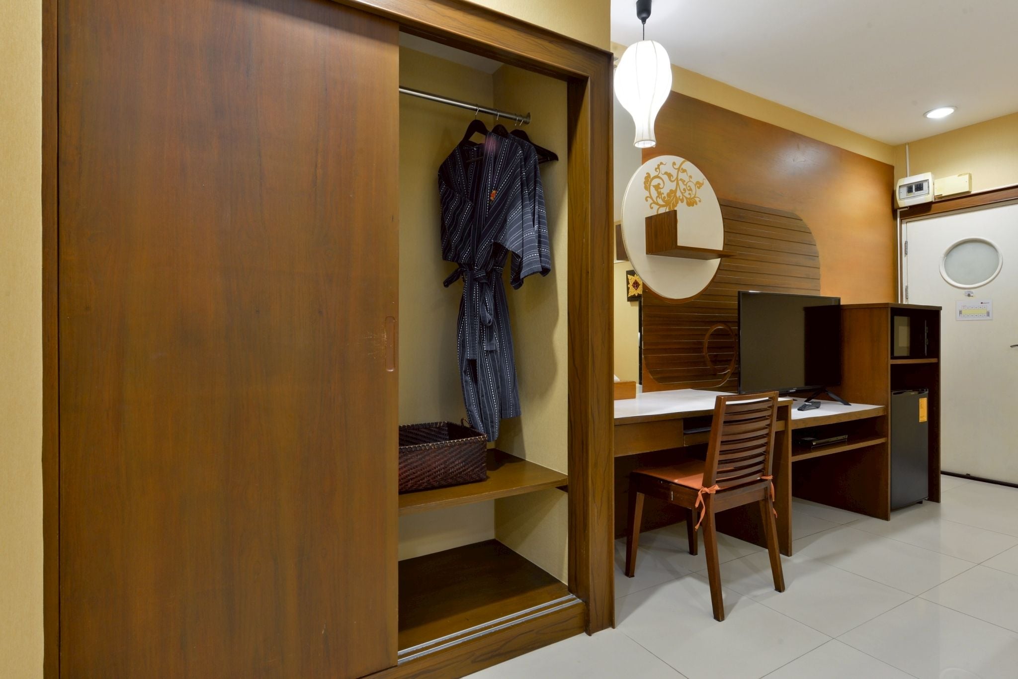 A compact room with a wooden wardrobe, a desk with a chair, a television, and a small refrigerator.