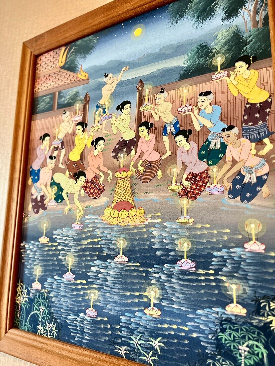 Un'opera d'arte incorniciata raffigura una scena tradizionale con persone in riva a un fiume, alcune delle quali tengono fiori di loto.
