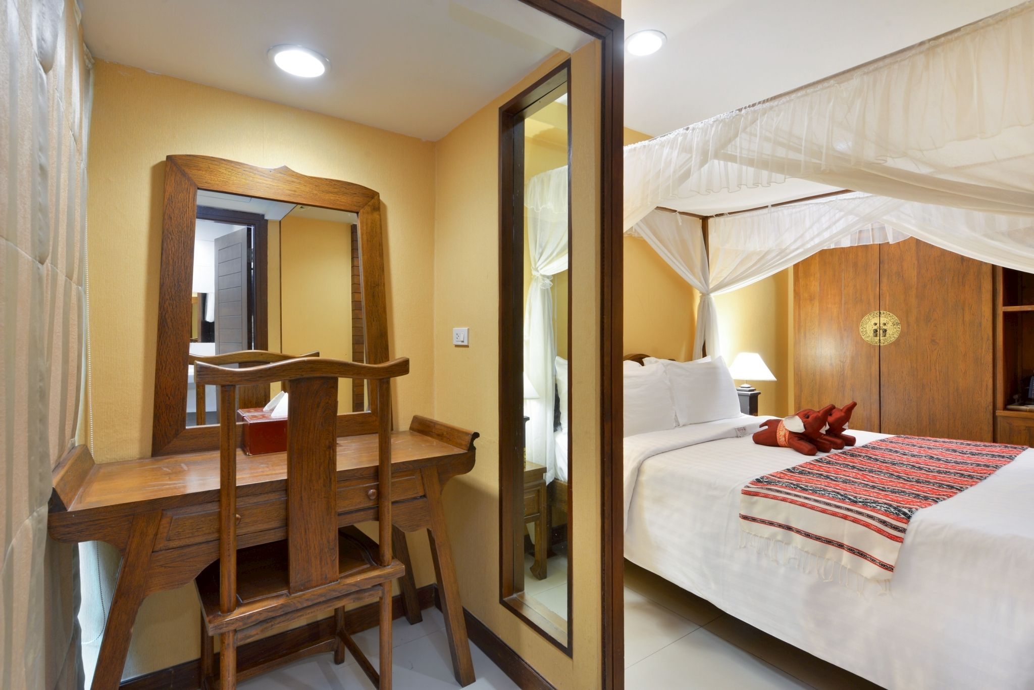 A hotel room with a wooden desk, a large mirror, a bed with a canopy, and a nightstand with a lamp.