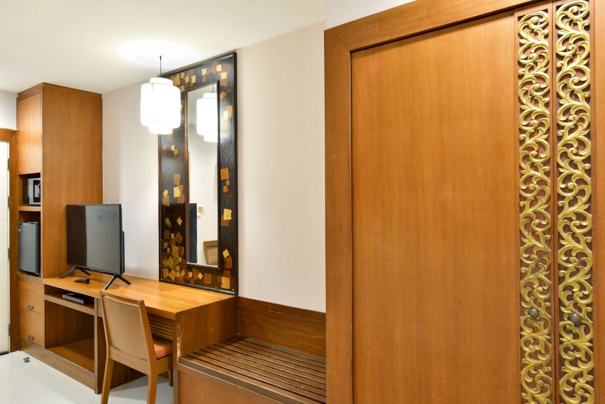 A hotel room with a wooden desk, a television, a chair, and a wardrobe with decorative panels.
