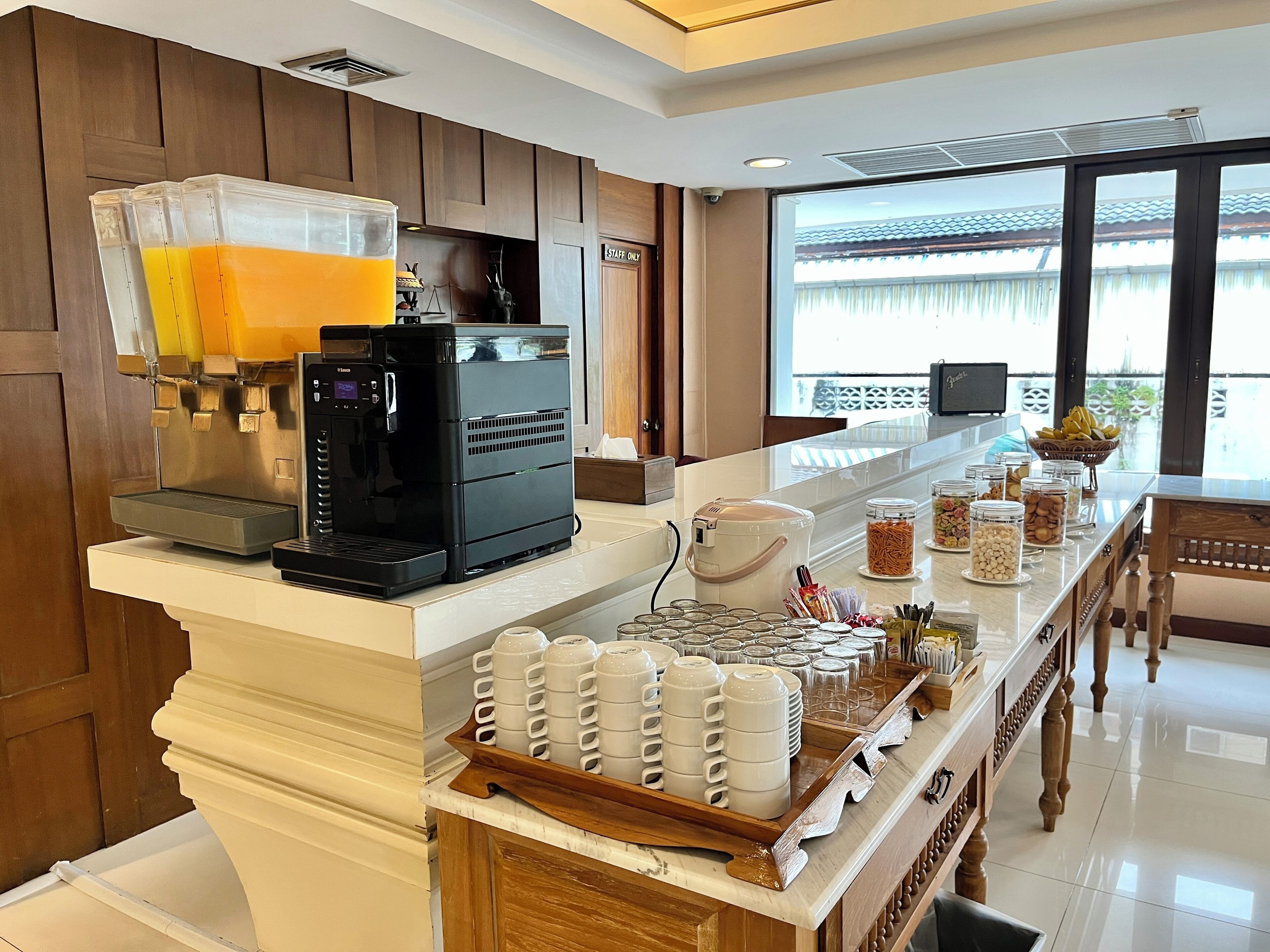 A hotel reception area with a coffee machine, a buffet counter with various food items, and a large window offering a view of the outside.