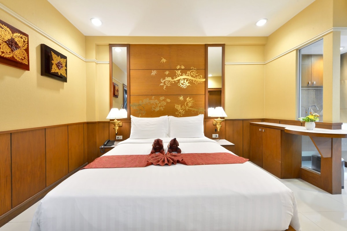 A hotel room with a large bed, two bedside lamps, a wooden headboard with floral carvings, a mirror, and a small desk with a vase of flowers.