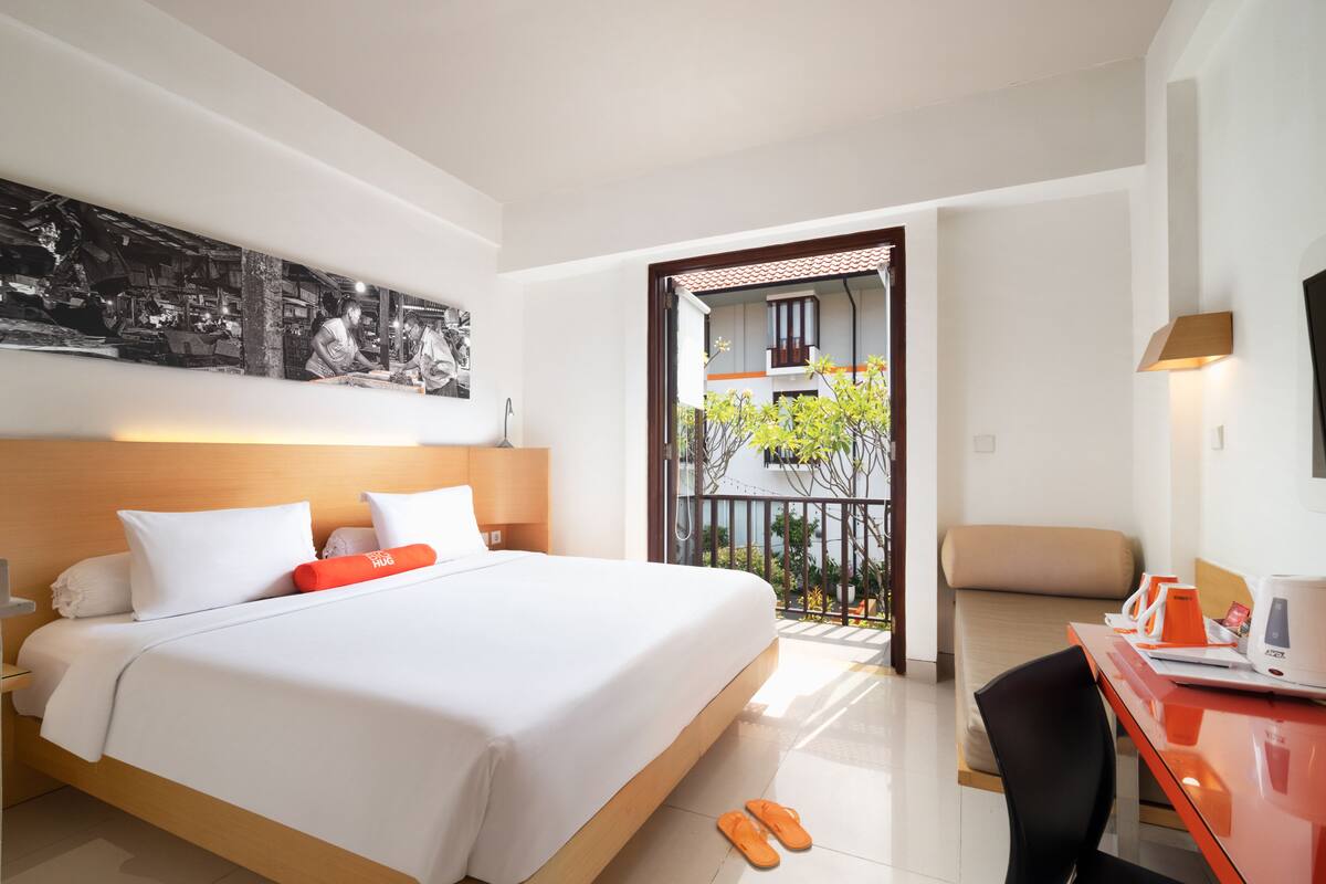 A hotel room with a large bed, a desk with a chair, and a balcony with a view of a building and greenery.