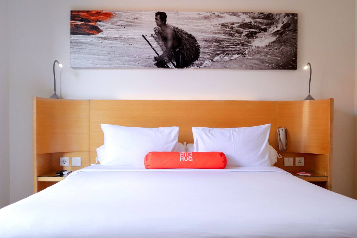 A hotel room with a large bed, white bedding, a wooden headboard, a red pillow, a wall-mounted painting, and bedside lamps.