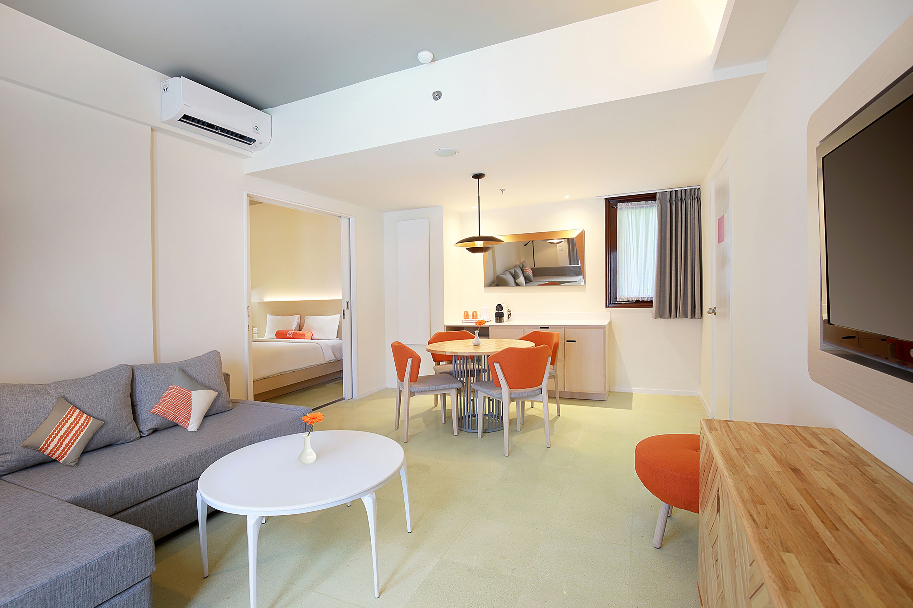 Quarto de hotel moderno com sofá cinzento, uma mesa redonda pequena, mesa de cabeceira em madeira, área de refeições com cadeiras laranja, uma cama e uma televisão.