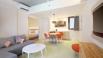 Quarto de hotel moderno com sofá cinzento, uma mesa redonda pequena, mesa de cabeceira em madeira, área de refeições com cadeiras laranja, uma cama e uma televisão.