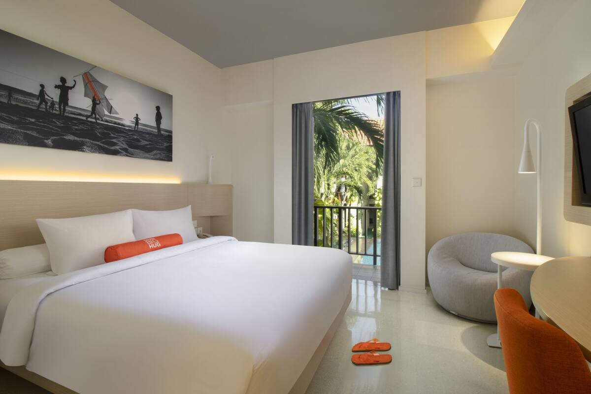 A modern hotel room with a large bed, a small round table, a chair, a TV, and a balcony with a view of palm trees.