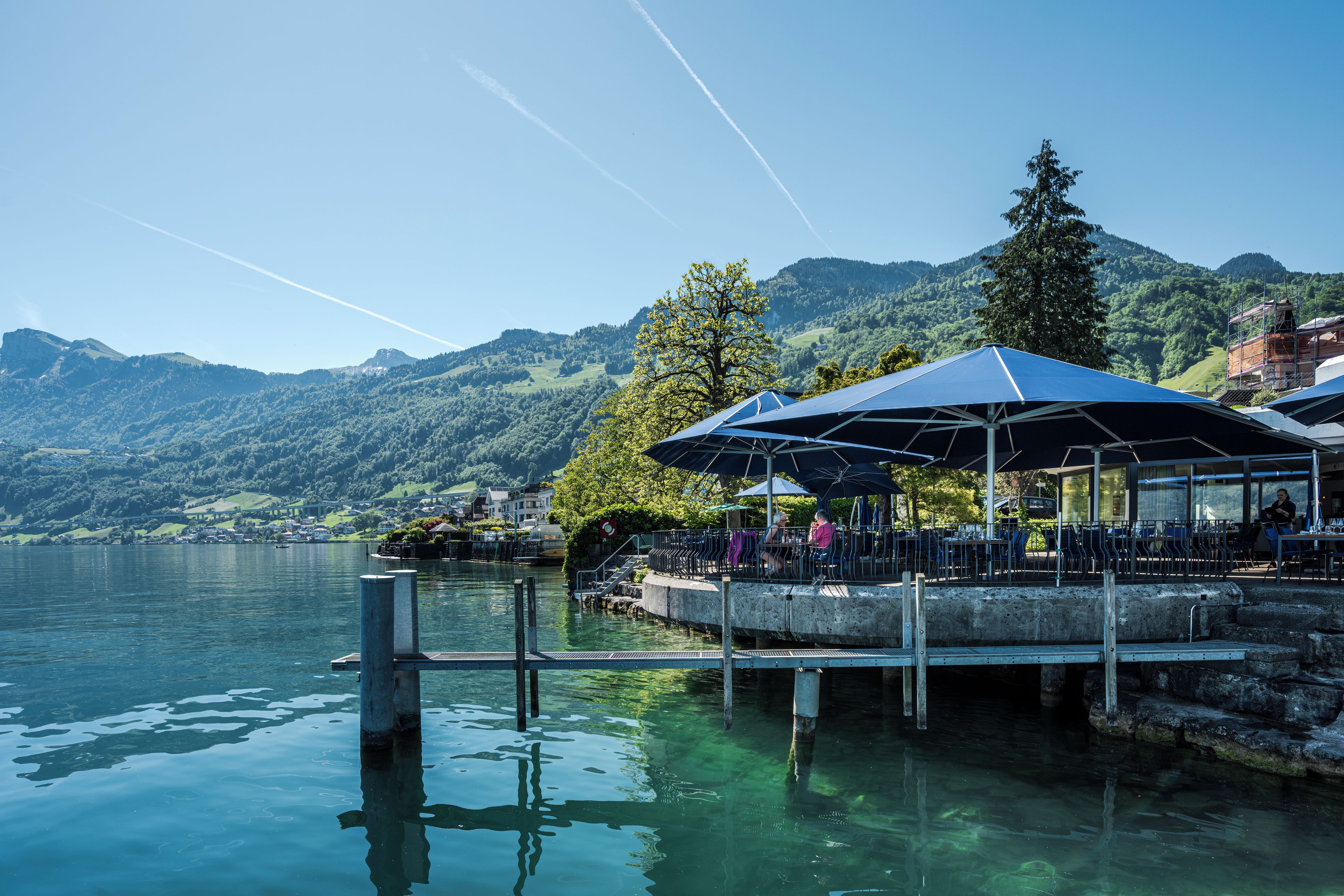 A lakeside restaurant with outdoor seating, a dock, and a mountain backdrop.