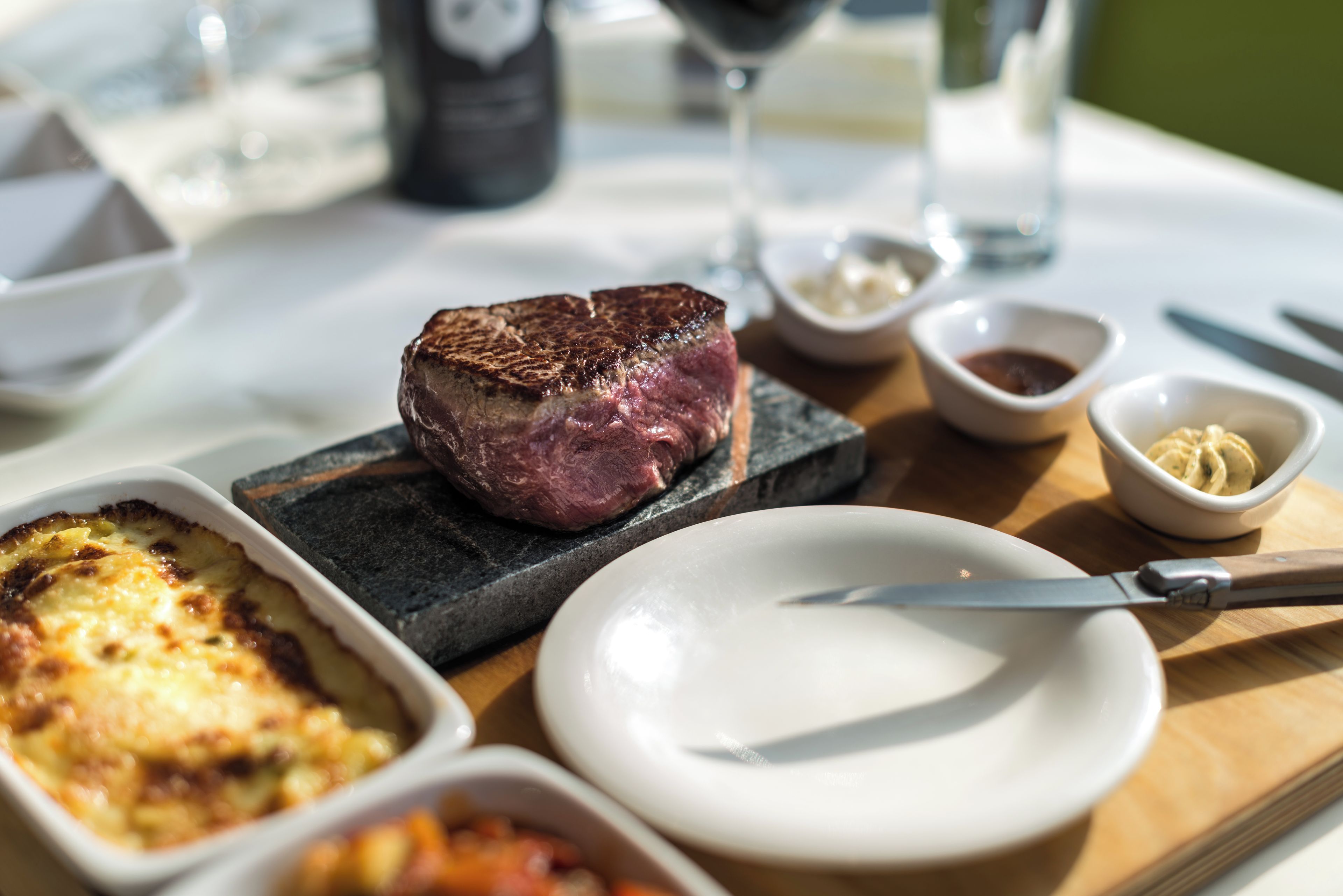 Un bistec sobre un plato de piedra con un cuchillo a un lado, un bol de salsa y un plato de patatas al horno.