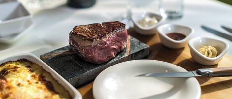 A steak on a stone plate with a knife beside it, a bowl of sauce, and a dish of baked potatoes.