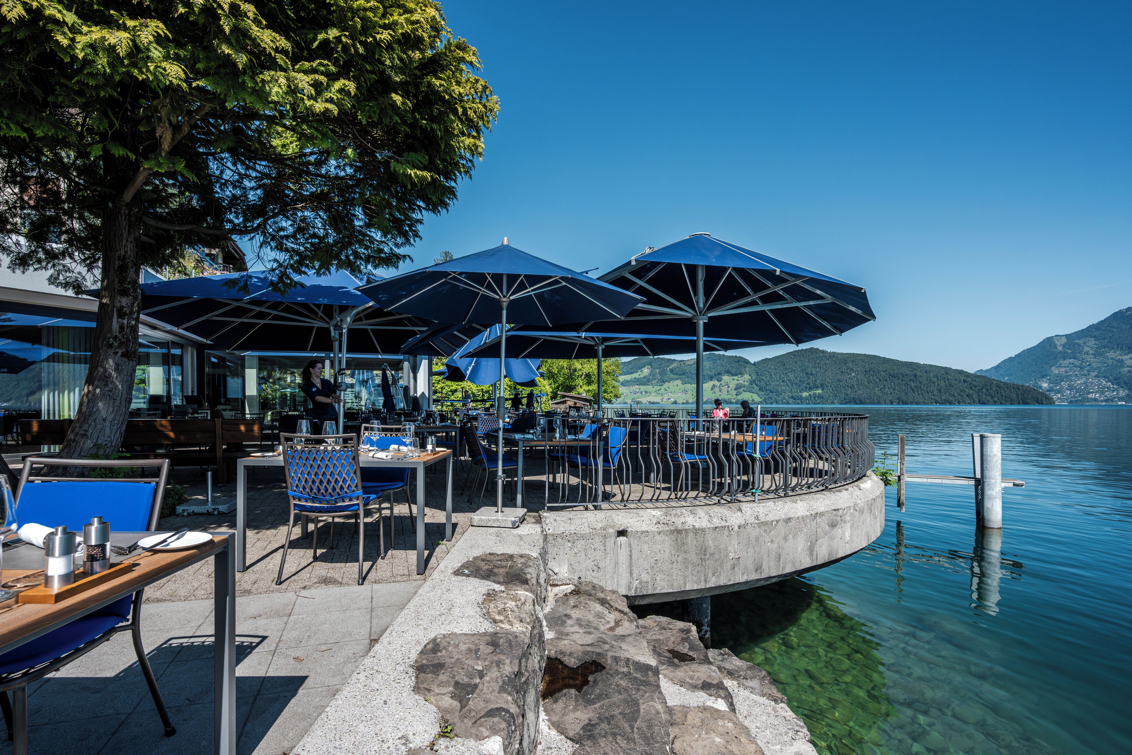 Un restaurante junto al lago con terraza, mesas y sombrillas azules.