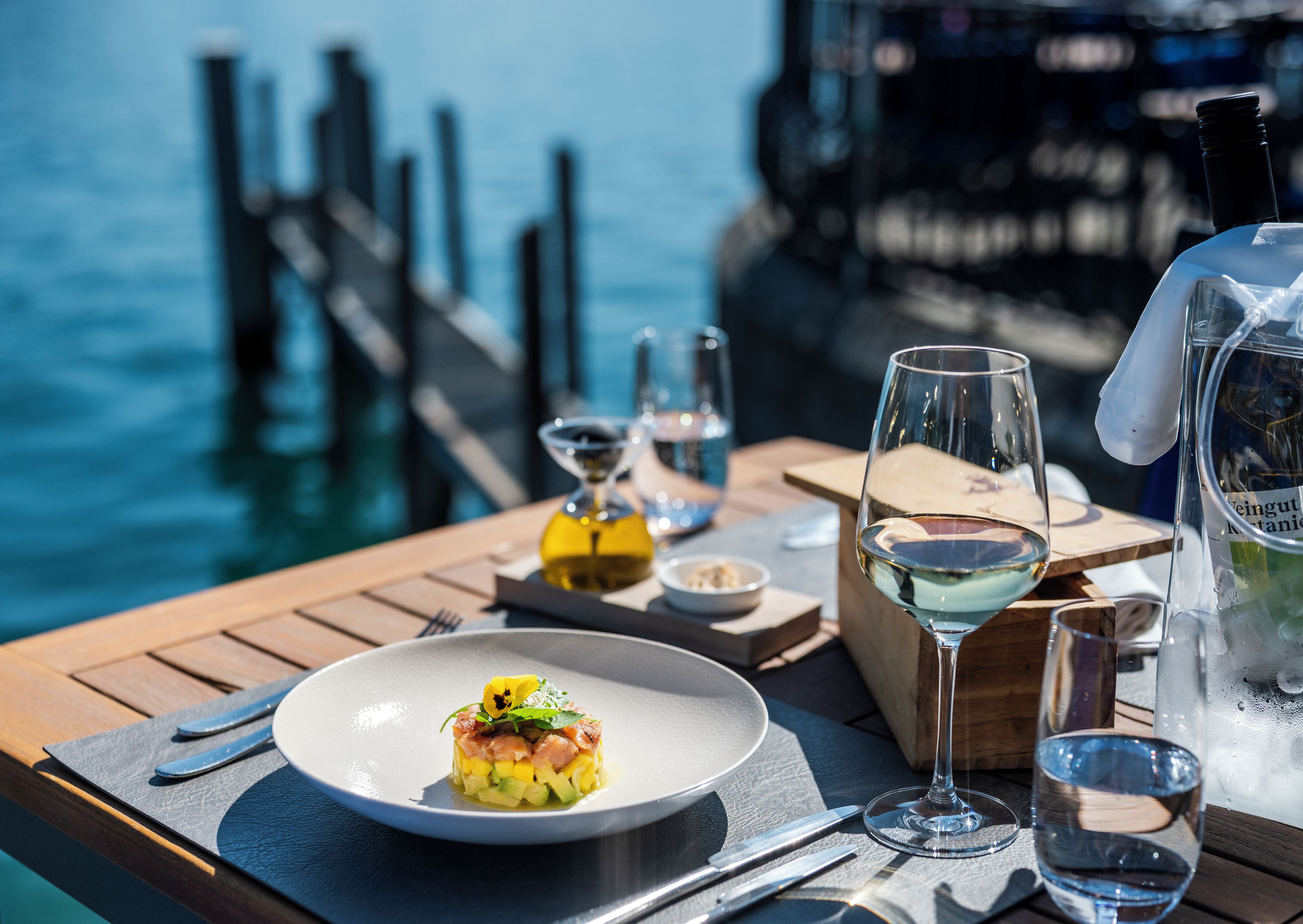Une table à manger avec une assiette de nourriture, des verres à vin et une carafe, le tout placé devant une jetée et l’eau.