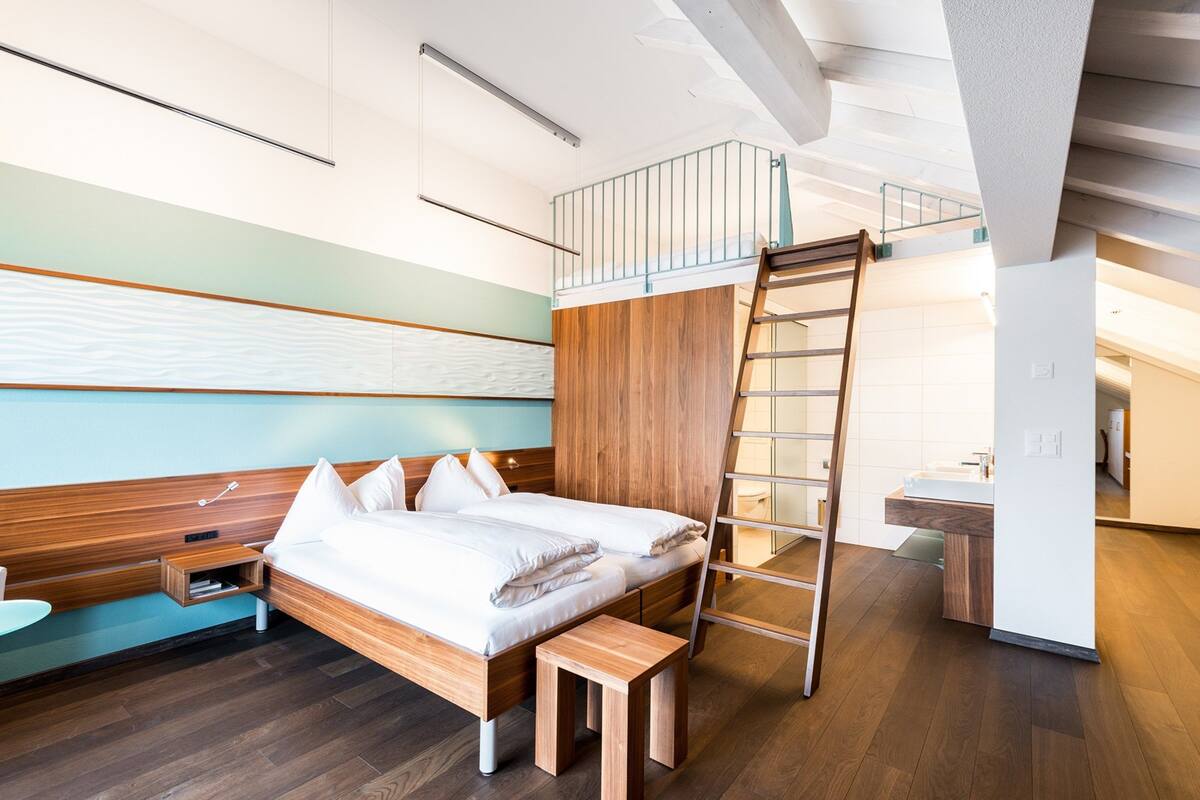 Habitación de hotel moderna con cama elevada, piso de madera, un escritorio pequeño y baño con lavamanos y espejo.