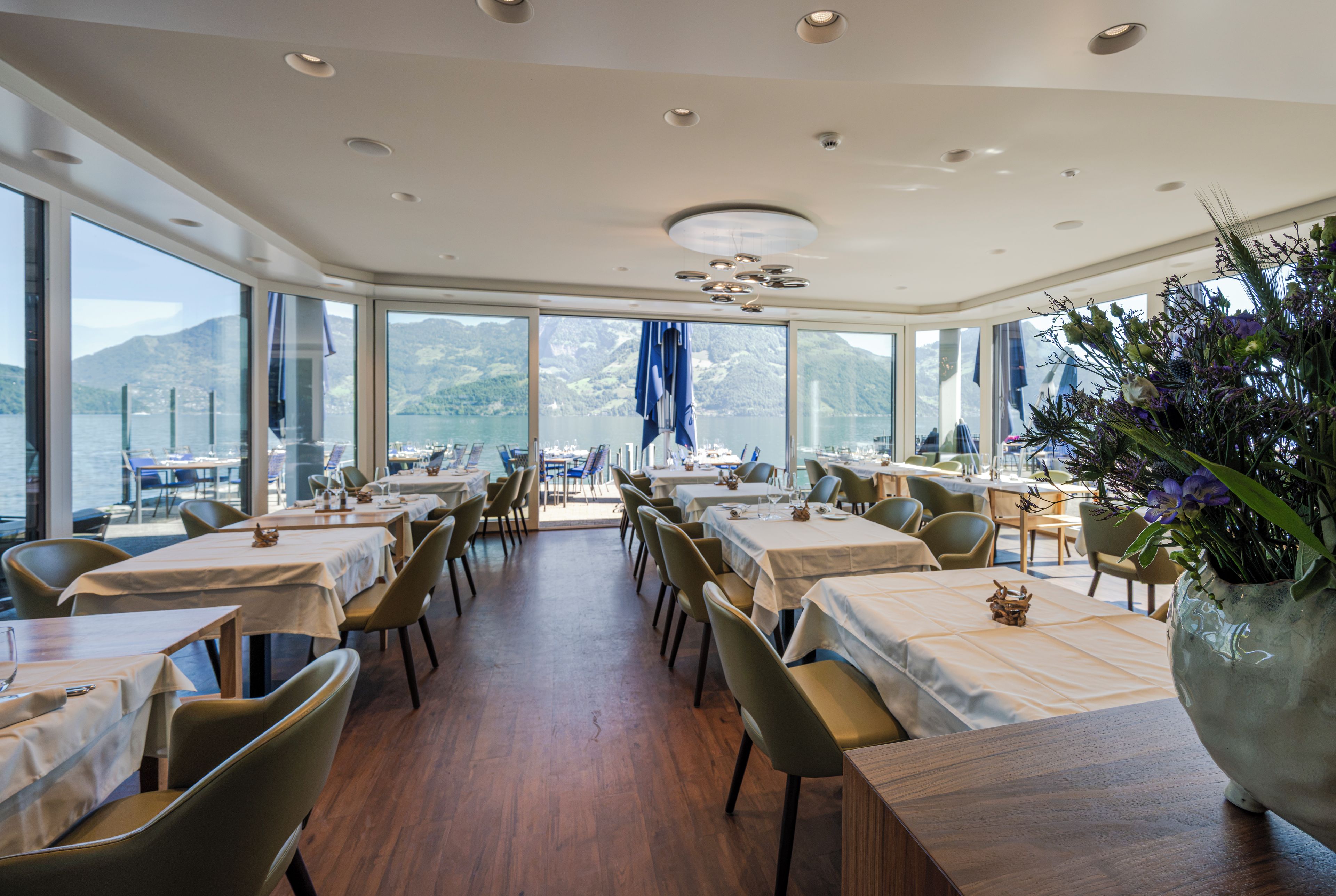 Ein Restaurant mit Tischen und Stühlen, große Fenster mit Blick auf die Berge und eine Decke mit Einbaustrahlern.