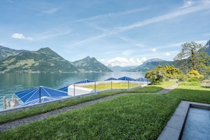 Vista del lago con zona verde, un muelle con sombrillas azules y montañas al fondo.