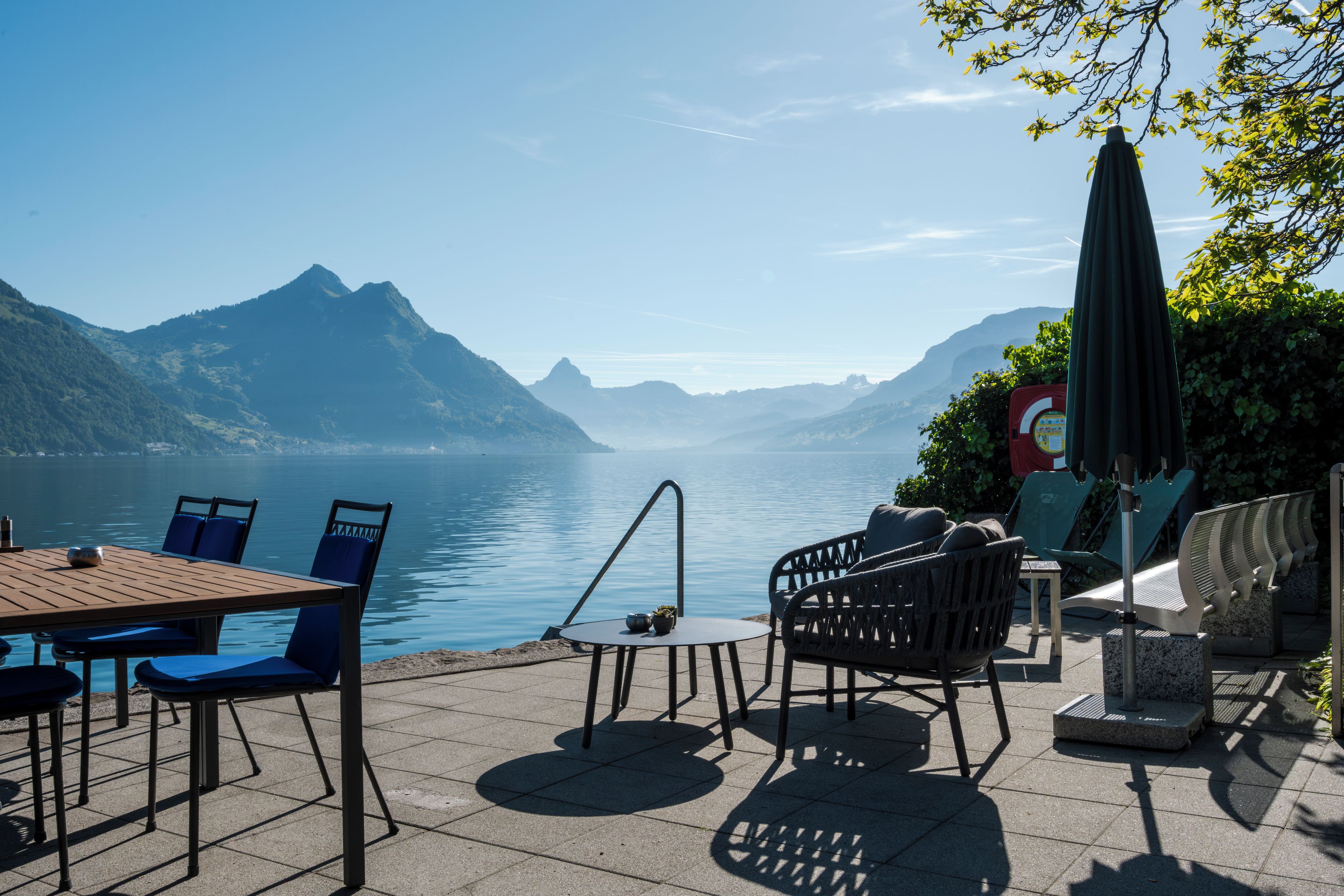 A terrace with outdoor seating, a table, and a view of a lake with mountains in the background.