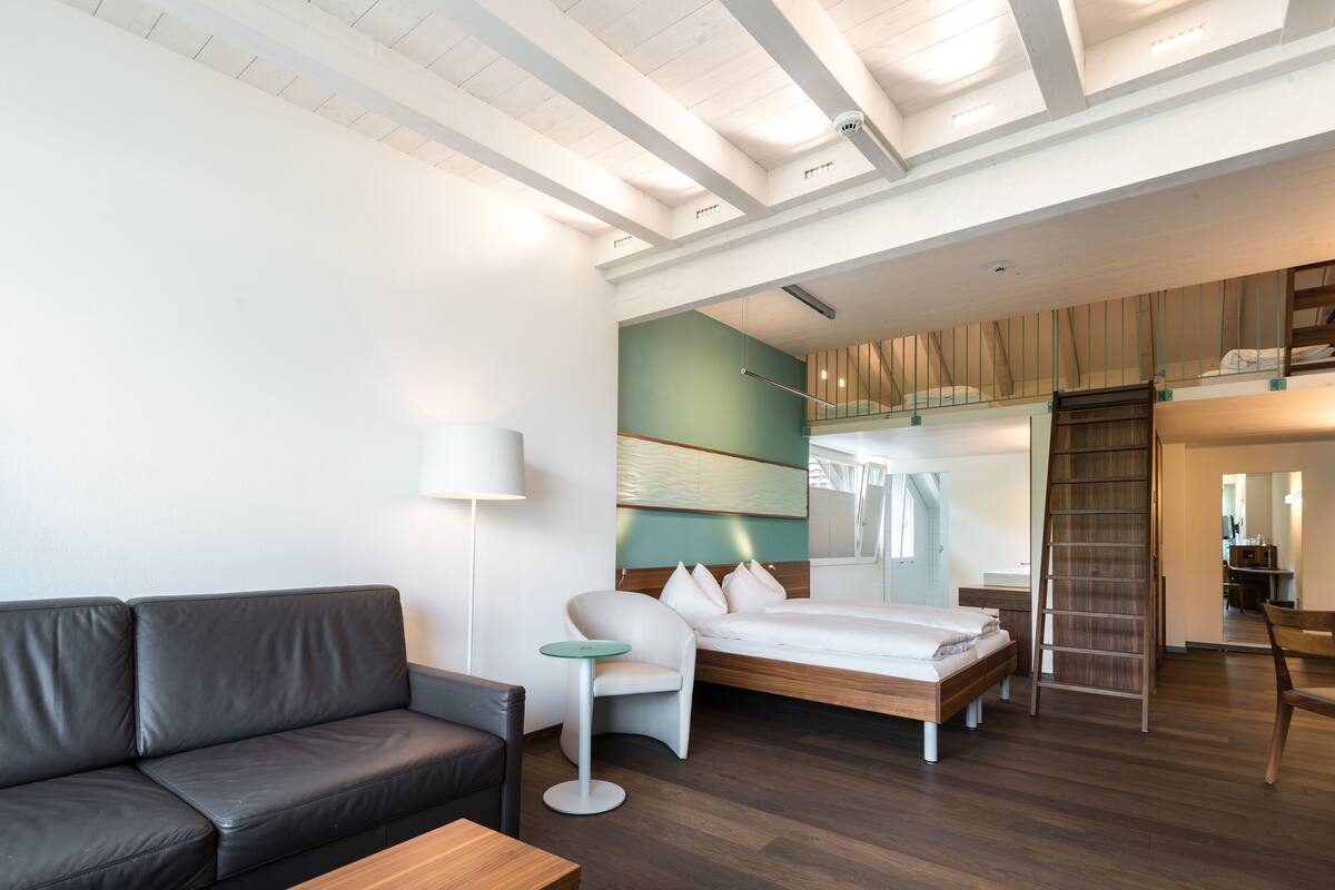 Una habitación de hotel moderna con sofá, una mesita redonda pequeña, cama con cabecero de madera y una escalera que lleva a un nivel superior.