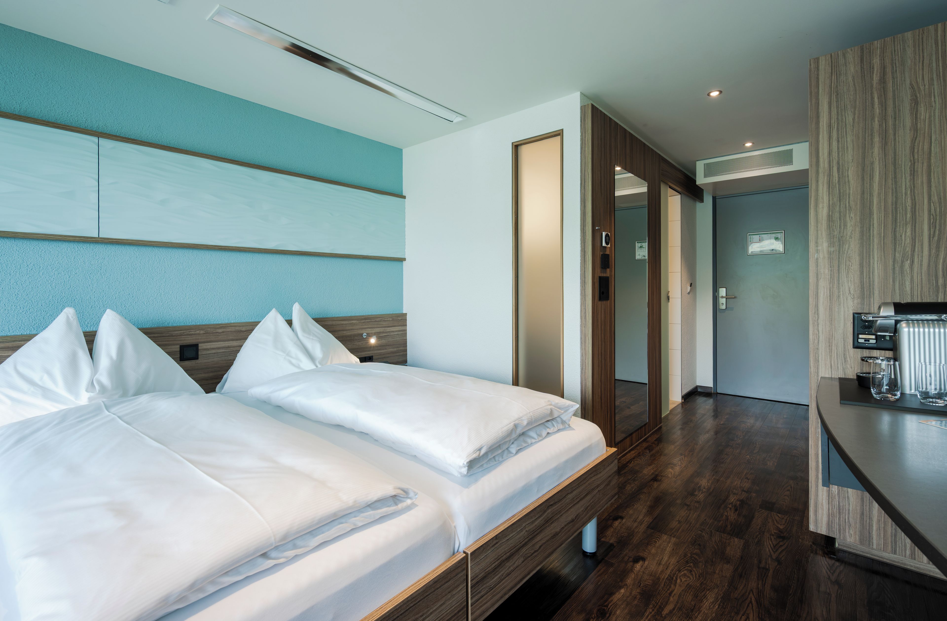 Ein Hotelzimmer mit zwei Betten, einem Holz-Kopfteil, einer blauen Akzentwand, Holzboden und einer Küchenzeile.
