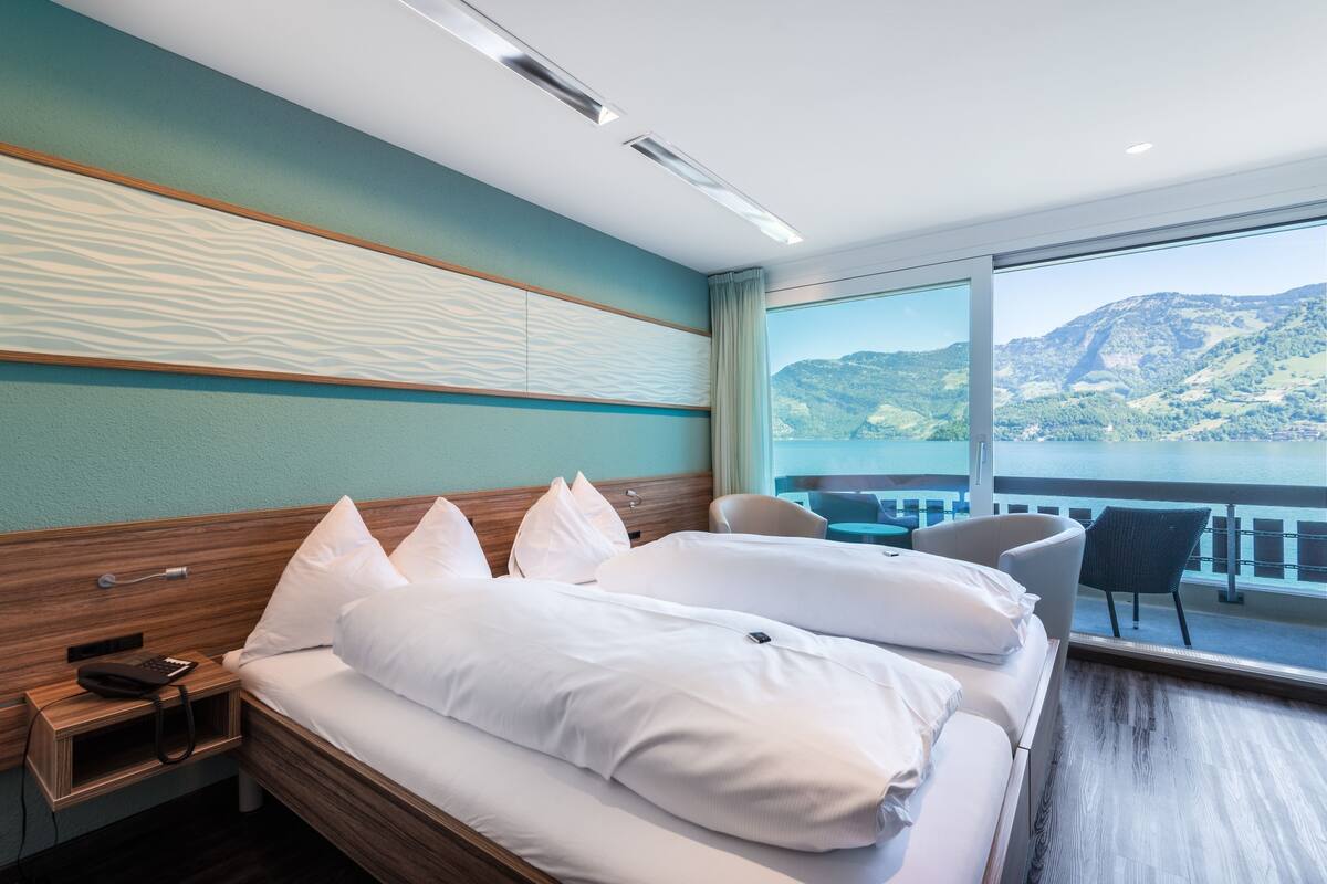Habitación de hotel con cama, mesita de noche, una silla y un amplio ventanal con vistas panorámicas a montañas y agua.