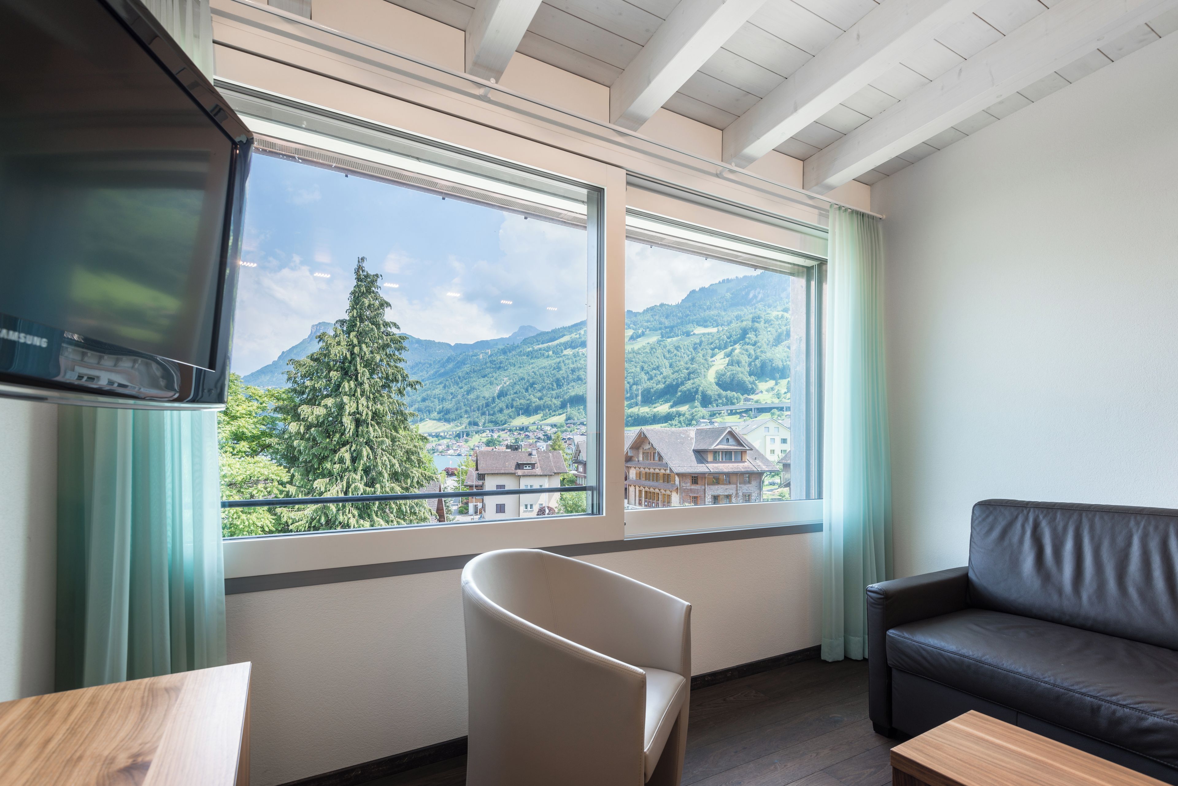 A room with a large window offering a view of a mountain and a village. There is a black leather sofa and a white chair.