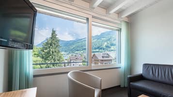 A room with a large window offering a view of a mountain and a village. There is a black leather sofa and a white chair.
