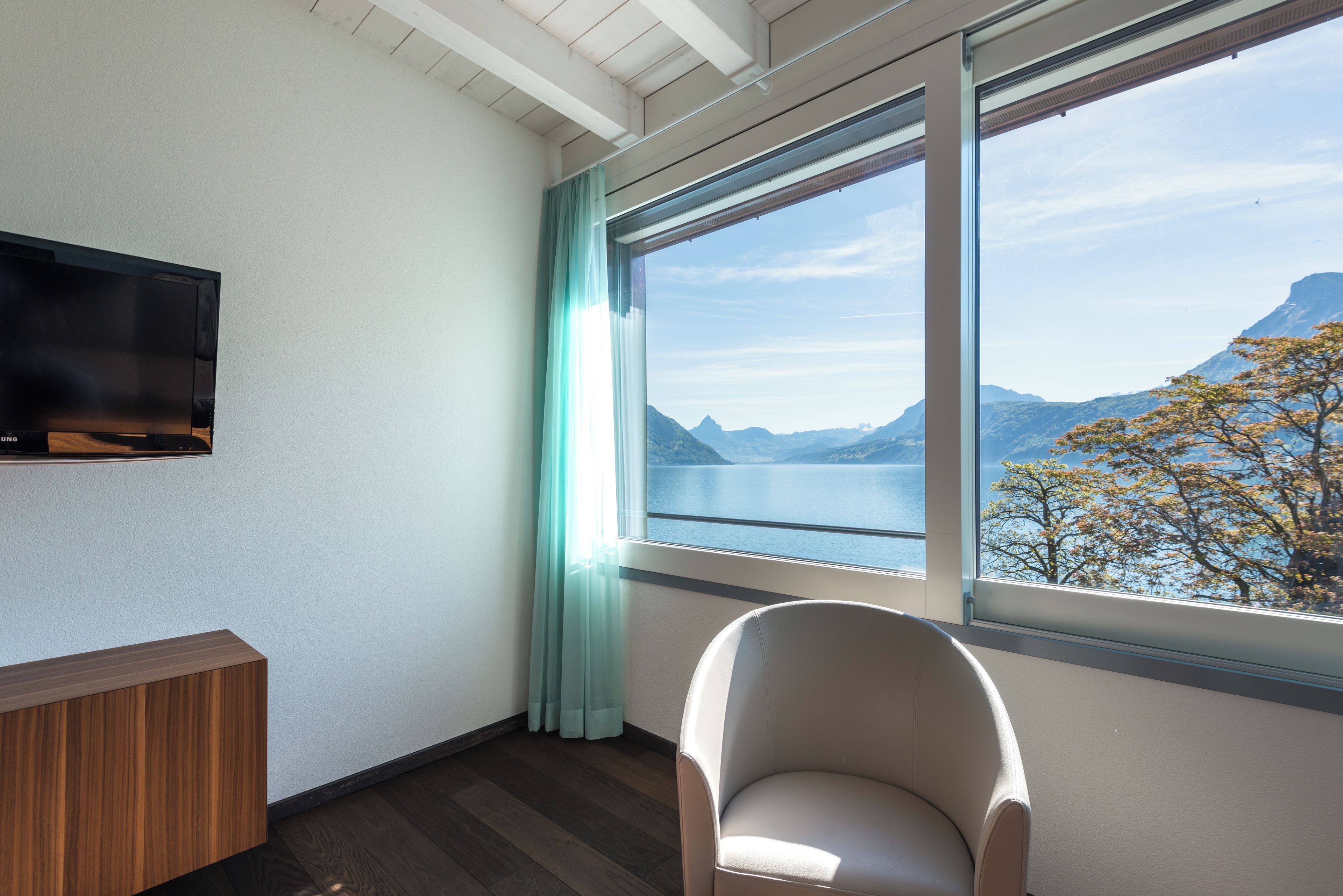 A room with a television mounted on the wall, a window with a view of mountains and a lake, and a chair near the window.