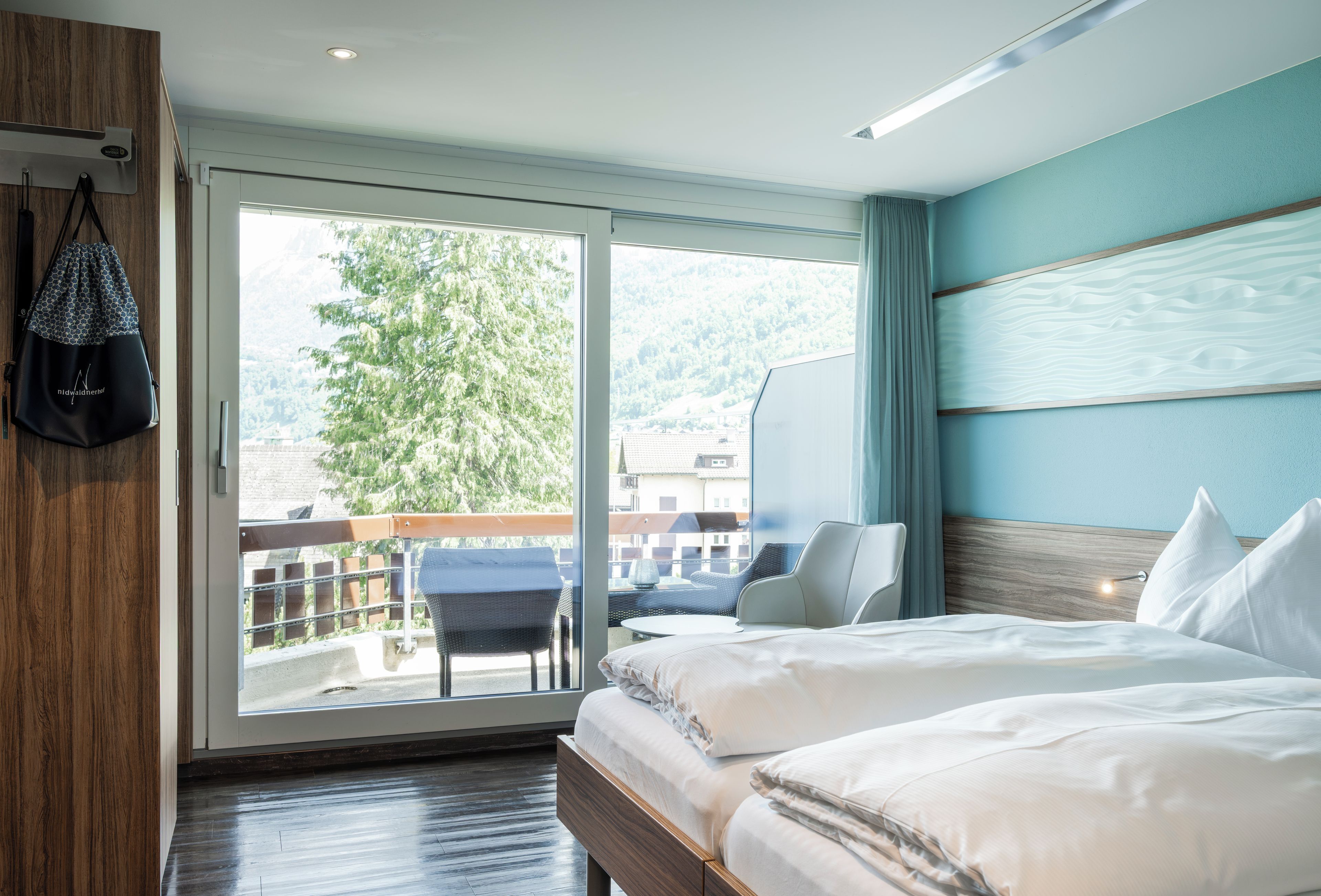 Ein Hotelzimmer mit Bett, Stuhl, Schreibtisch und Blick auf einen Balkon sowie Bäume.