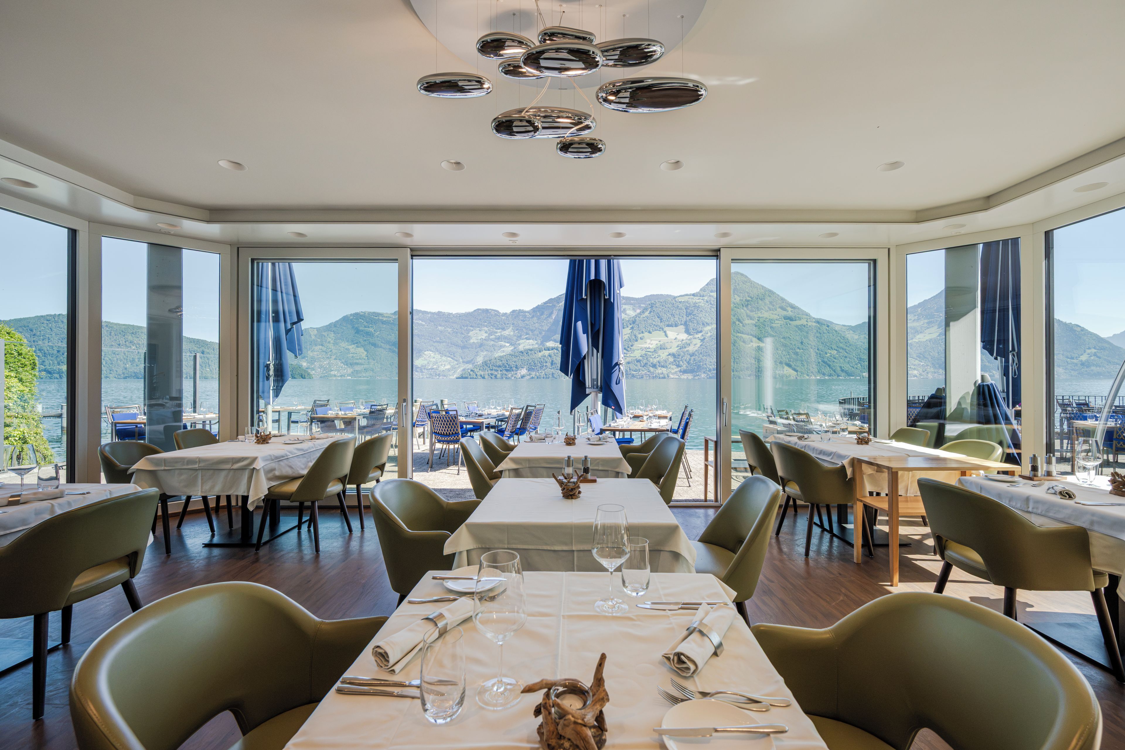 A restaurant with tables set for dining, large windows offering a view of the water and mountains, and a modern ceiling design.