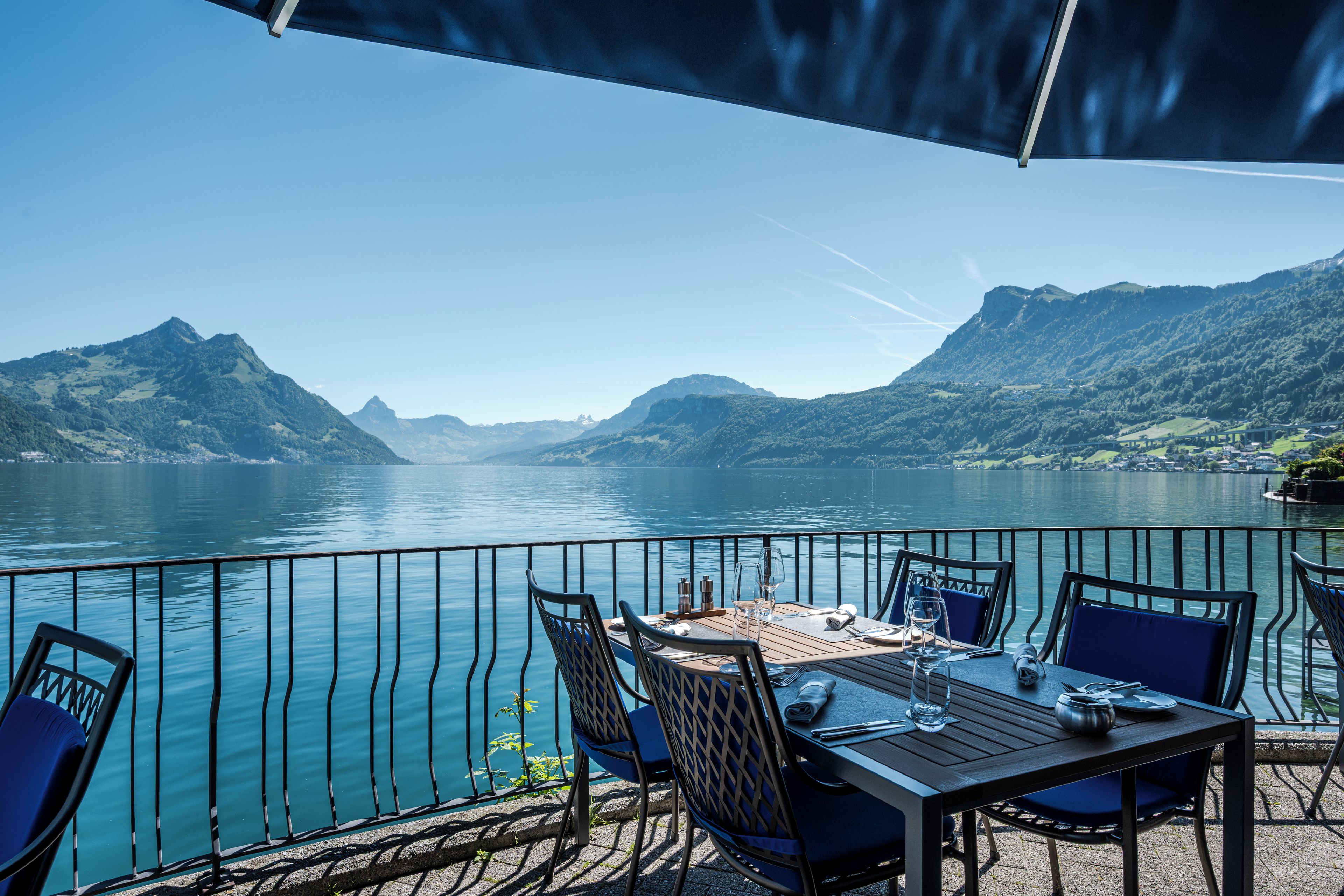 An outdoor dining area with a table and chairs, overlooking a lake with mountains in the background.