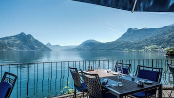 An outdoor dining area with a table and chairs, overlooking a lake with mountains in the background.