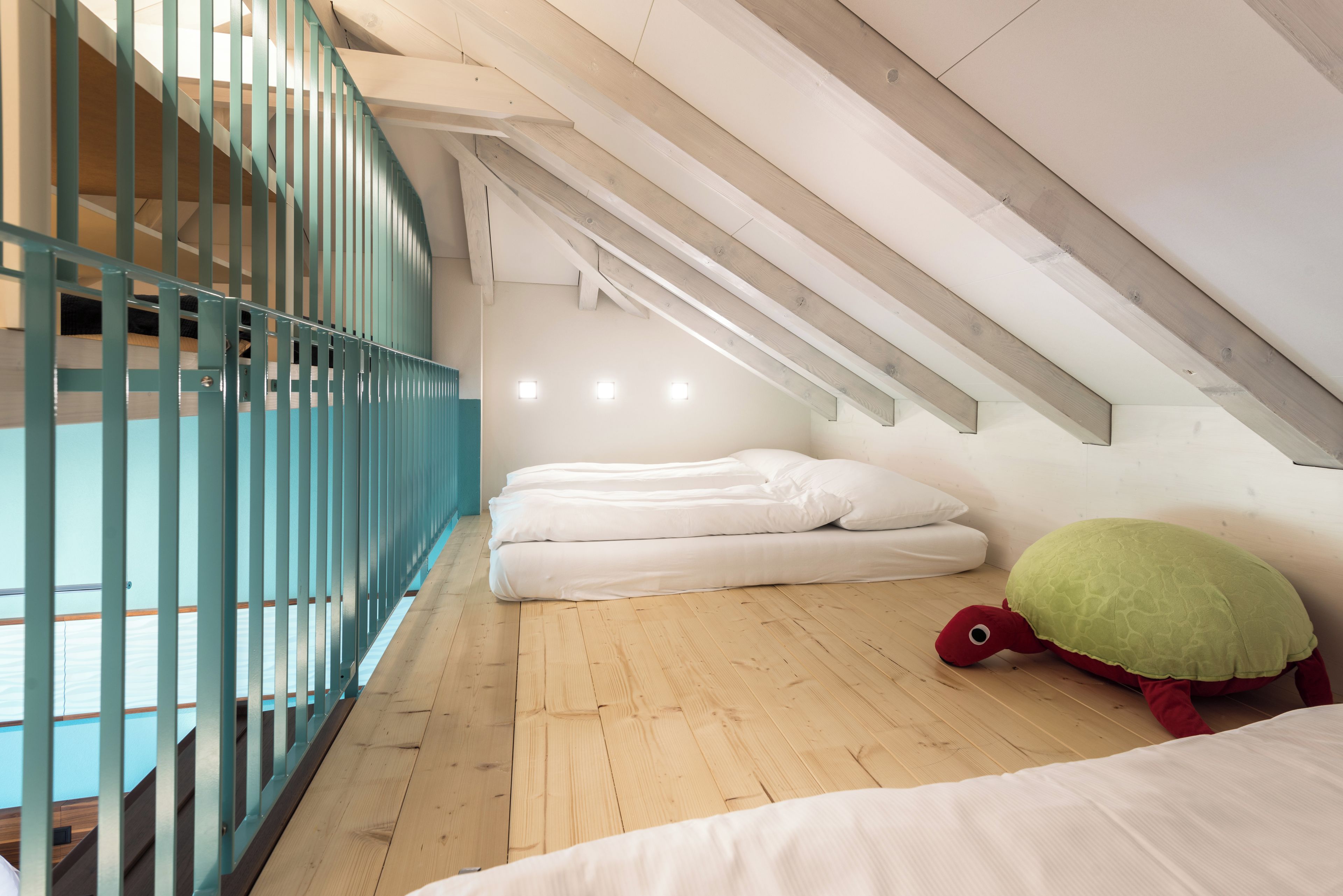A room with a sloped ceiling, wooden flooring, a bed with white bedding, a green turtle plush toy, and a blue railing.