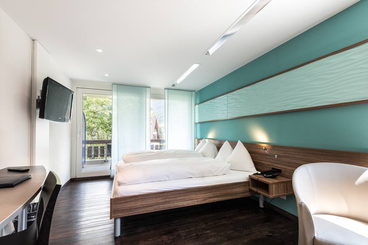 Habitación de hotel moderna con cama, escritorio, silla, televisor y balcón con vistas a zonas verdes.
