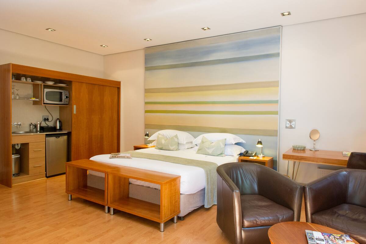 A hotel room with a bed, a desk, a chair, a kitchenette, and a large painting on the wall.