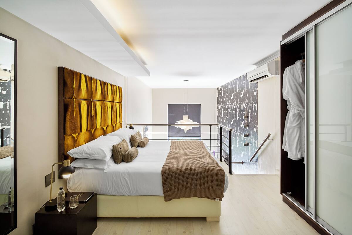 A modern bedroom with a large bed, a headboard, a bedside table with a lamp, and a view of the city through a balcony.