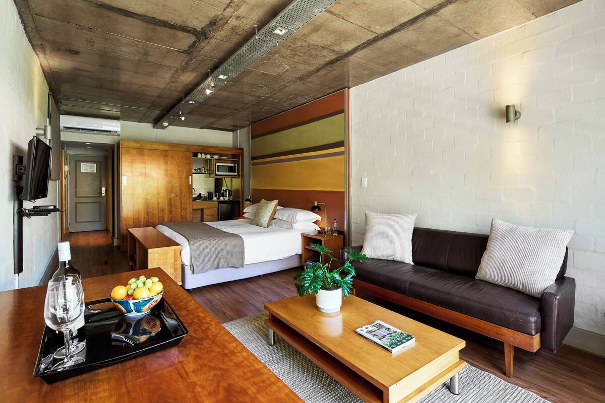 A modern hotel room with a bed, a sofa, a wooden coffee table, and a small kitchen area.