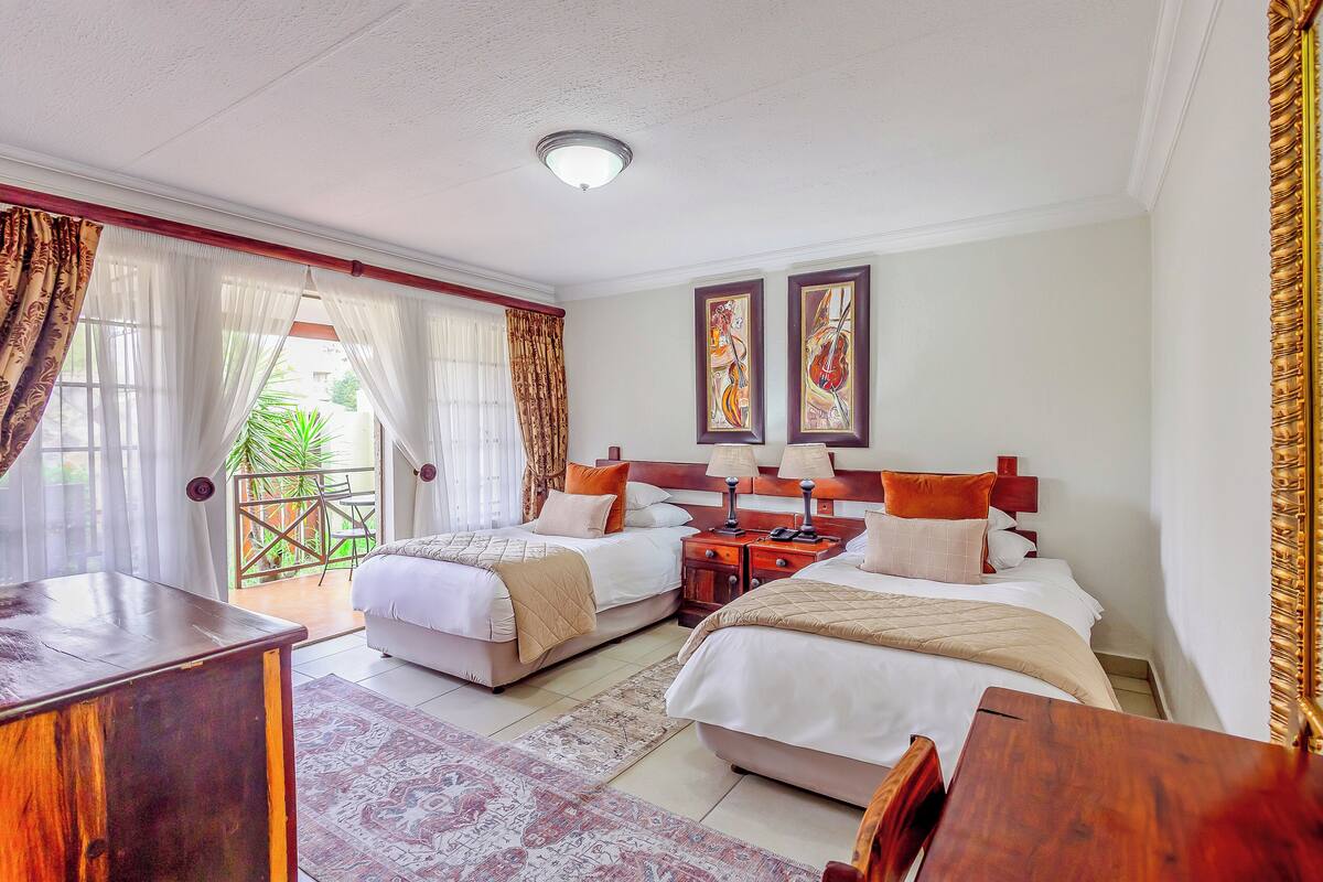 A hotel room with two beds, a wooden desk, a nightstand, and a balcony with a view of greenery.