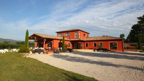 A two-story building with a red facade and a tiled roof, surrounded by a gravel driveway and a grassy area with white lounge chairs.