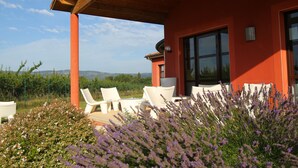A covered outdoor seating area with white chairs, a red building, and a view of the landscape.