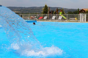 A swimming pool with a person swimming, a water slide, and a fenced area with lounge chairs. - Vacancéole - Résidence le Domaine du Lac (Chateauneuf-sur-Isere)