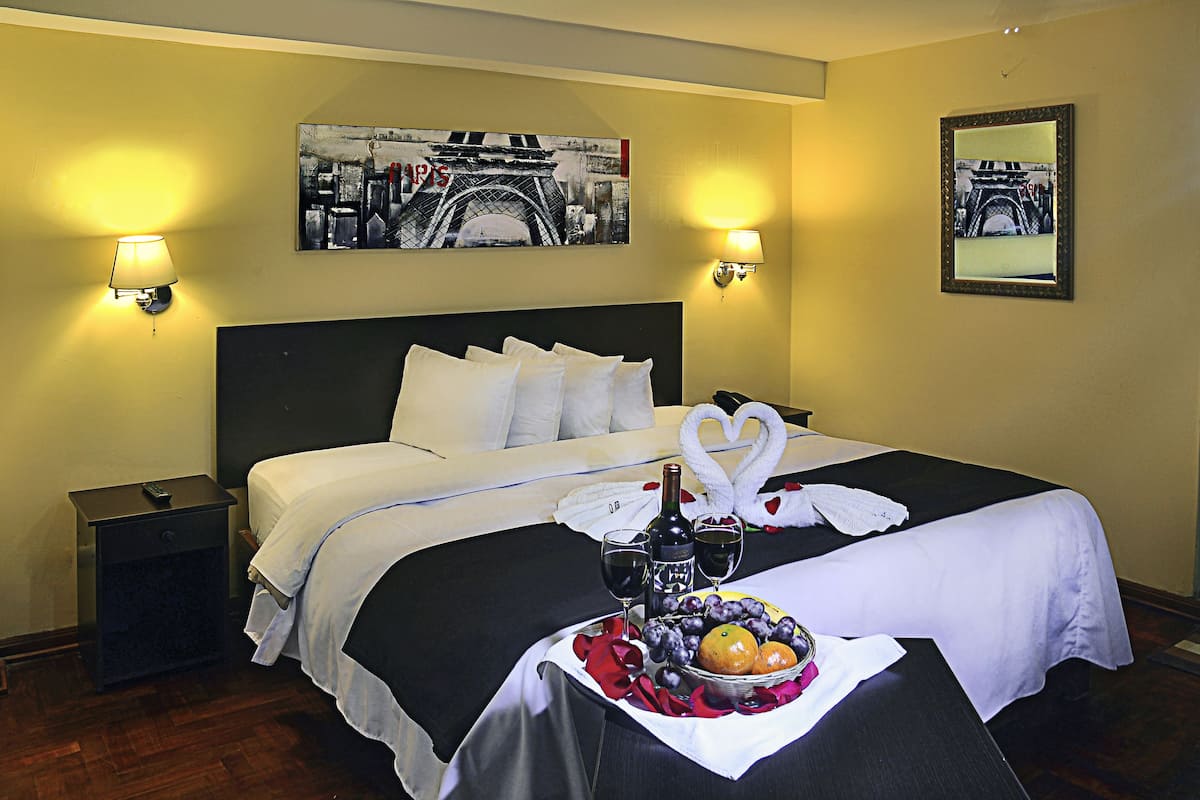 A hotel room with a bed, two wall-mounted lamps, a painting of the Eiffel Tower, a mirror, and a tray with fruits and wine glasses.
