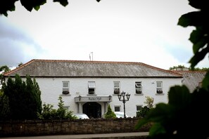 A white building with a sign reading "Racket Hall Hotel." It has a tiled roof, multiple windows, and a stone wall in front.